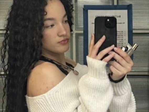 Karma holds a phone up to take a mirror selfie. Her black hair falls behind her back and she wears a white sweater
