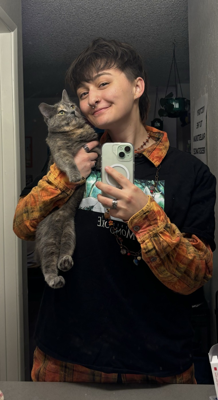 Photo of a white person with short dark  hair, holding up a grey cat and taking a selfie in a mirror