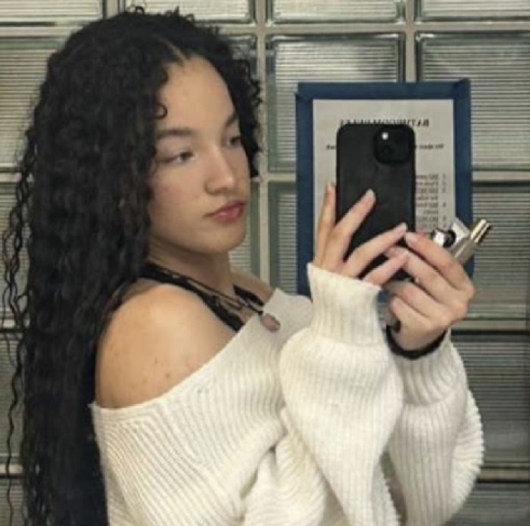 Karma holds a phone up to take a mirror selfie. Her black hair falls behind her back and she wears a white sweater