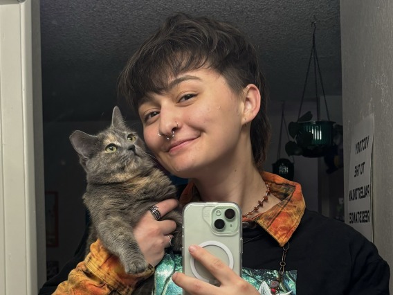 Photo of a white person with short dark  hair, holding up a grey cat and taking a selfie in a mirror