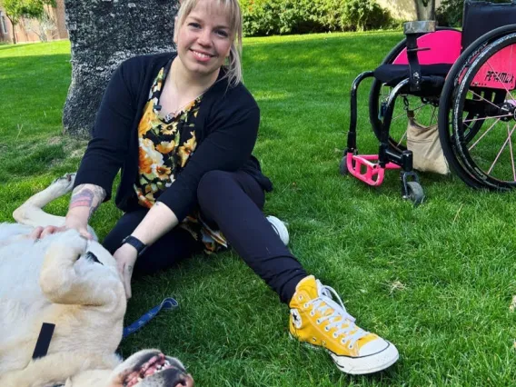 a photo of Sarah Heinzl, who sits in the grass beside her pink and black wheelchair. She is petting her dog Aiden, who is rolling in the grass