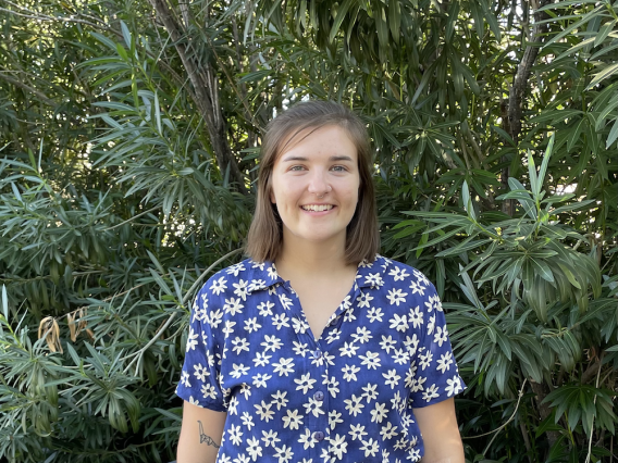 Picture of Hannah Miller. Hannah is wearing a blue button shirt and is featured smiling against natural, green background