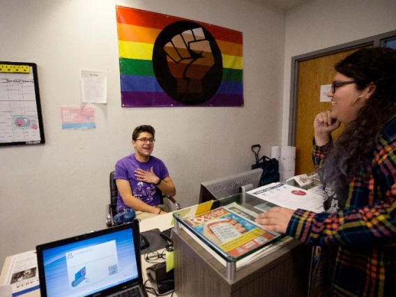 Two members of LGBTQ conversing at a desk, a pride flag hanging behind them on the wall