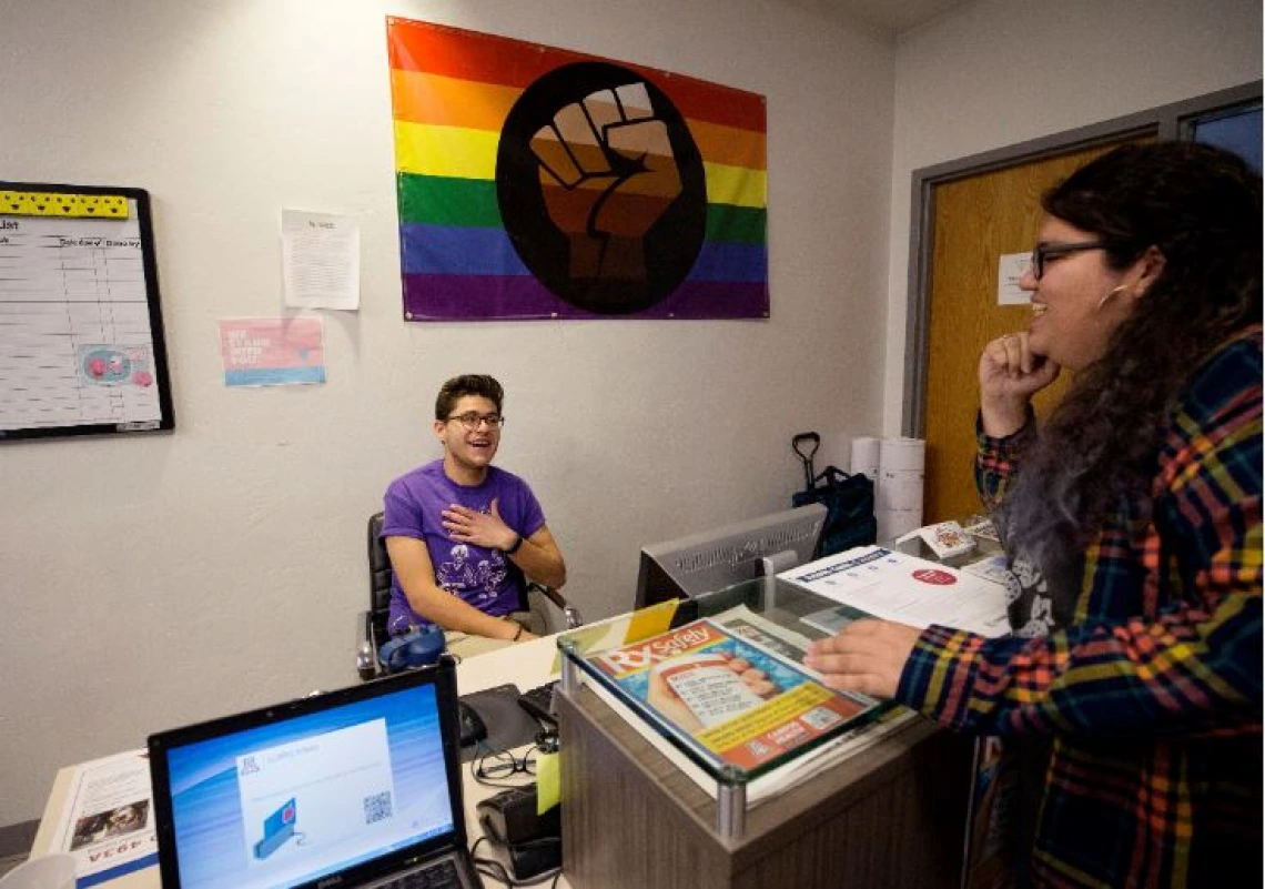 Two members of LGBTQ conversing at a desk, a pride flag hanging behind them on the wall