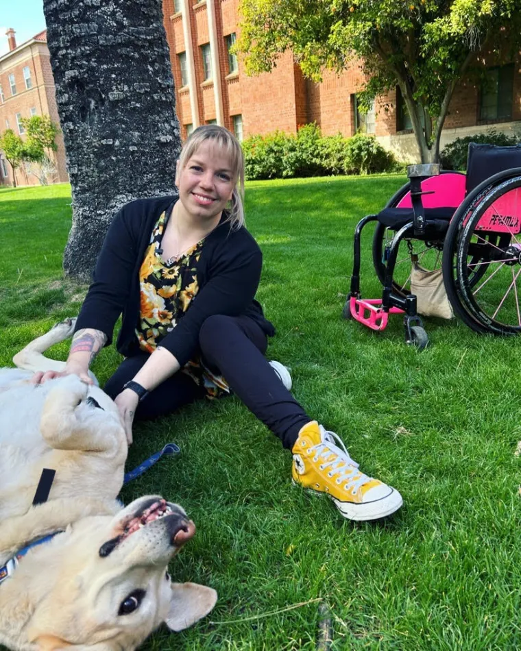 a photo of Sarah Heinzl, who sits in the grass beside her pink and black wheelchair. She is petting her dog Aiden, who is rolling in the grass